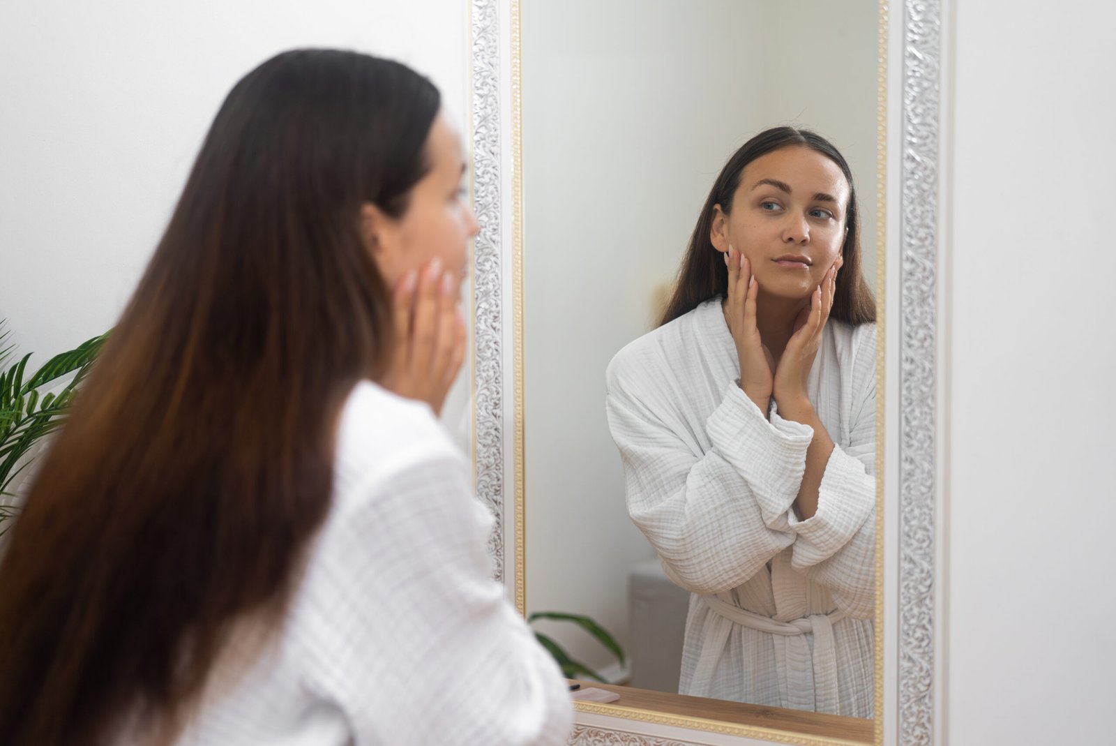Attractive brunette woman in gown looking in mirror in bathroom lady checking facial skin condition standing by dressing table at home woman with healthy skin near mirror