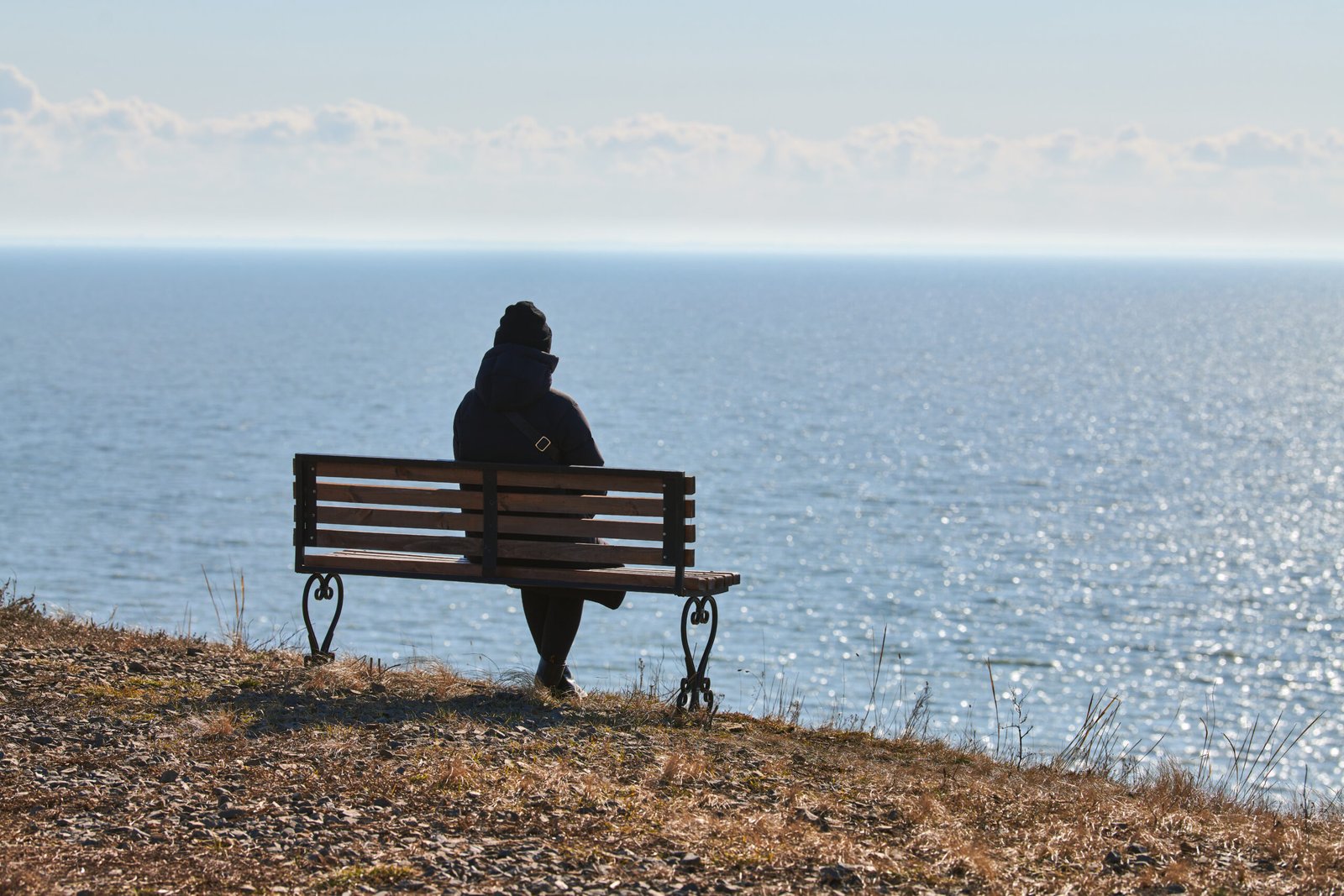 Single girl in a black jacket and hat sitting on bench at cliff at front of sea, peaceful and quiet place for thinking alone, loneliness and loss of loved one concept. Pacifying view of marine horizon