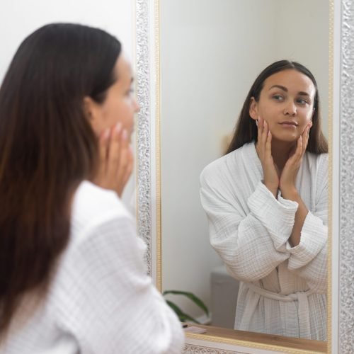 Attractive brunette woman in gown looking in mirror in bathroom lady checking facial skin condition standing by dressing table at home woman with healthy skin near mirror