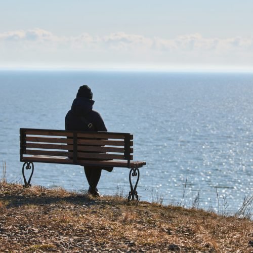 Single girl in a black jacket and hat sitting on bench at cliff at front of sea, peaceful and quiet place for thinking alone, loneliness and loss of loved one concept. Pacifying view of marine horizon