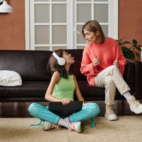 Teenage girl sitting on the floor with laptop and teasing her mother while she talking seriously with her in the room at home