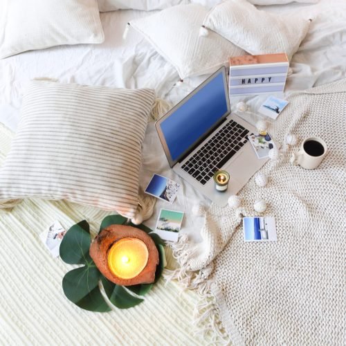 From above various instant photos from summer vacation and mug of hot beverage placed on soft bed near laptop with blue screen at home